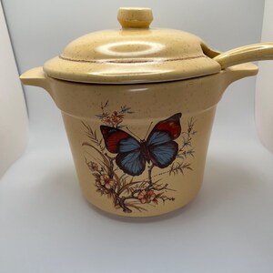 May include: A beige ceramic soup tureen with a lid and ladle. The tureen features a butterfly and floral design in red, blue, and brown. The lid has a small knob and a brown speckled pattern.