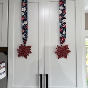 May include: Two white kitchen cabinet doors with black handles are decorated with Christmas poinsettia ribbon hangers. Each hanger has a dark blue ribbon with red and white floral patterns, and a red poinsettia ornament.