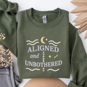 May include: Olive green sweatshirt with the words "ALIGNED and UNBOTHERED" in a whimsical font. The design includes a crescent moon, stars, and wavy lines. The sweatshirt is made of a soft material.