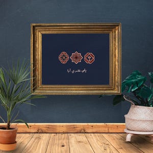 Puede incluir: Una obra de arte enmarcada con un marco dorado. La obra presenta tres diseños geométricos en rojo y blanco sobre un fondo azul oscuro. El texto debajo de los diseños está en un idioma extranjero.