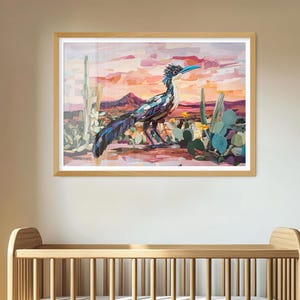 May include: A framed wall art print featuring a stylized roadrunner bird standing in a desert landscape with cacti and succulents. The background displays a sunset with shades of pink, orange, and purple, set above a wooden baby crib.