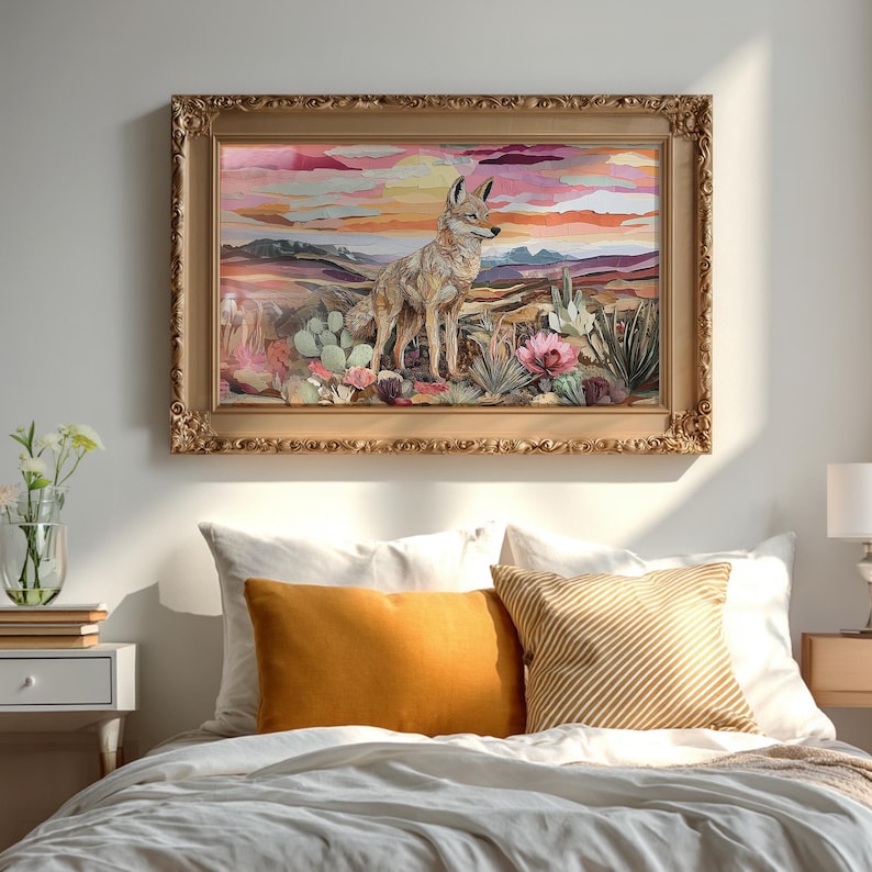 May include: A framed artwork depicts a coyote in a desert landscape with cacti and flowers under a colorful sunset. The ornate gold frame complements the warm tones of the artwork. The image hangs above a bed with orange and striped pillows.