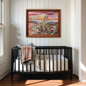 May include: A framed artwork depicting a coyote in a desert landscape with cacti and flowers, set against a sunset sky. The artwork hangs above a black crib with bedding and a blanket.