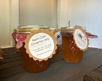 May include: Glass jars of amber-colored Apple Cider Cinnamon Jelly with gold metal lids. Each jar features a red patterned fabric topper, a scalloped brown paper tag with text, and a secondary round tag with the Cotton and Clay Farm Stand logo.
