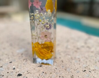 May include: A clear rectangular resin block with pressed flowers and petals. The flowers are pink, white, and yellow. The block is sitting on a textured surface.