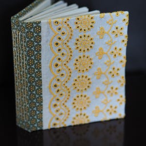May include: Open journal with a light blue cover featuring yellow floral embroidery. The spine is dark green with a repeating yellow floral pattern. The pages are white and the journal is open, displaying the interior.