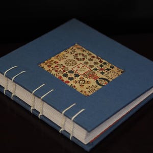 May include: A handmade journal with a blue cover and a decorative patterned insert. The journal is bound with white thread and has a rectangular design on the front cover. The pages are white with red edges.
