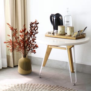 May include: A brown vase with a textured surface holds a bouquet of dried red leaves. The vase is on a light brown rug in front of a white table with a wooden top and white legs. On the table is a black electric kettle, two mugs, a glass bottle with the word "vibe" on it, and a wooden tray with various items.