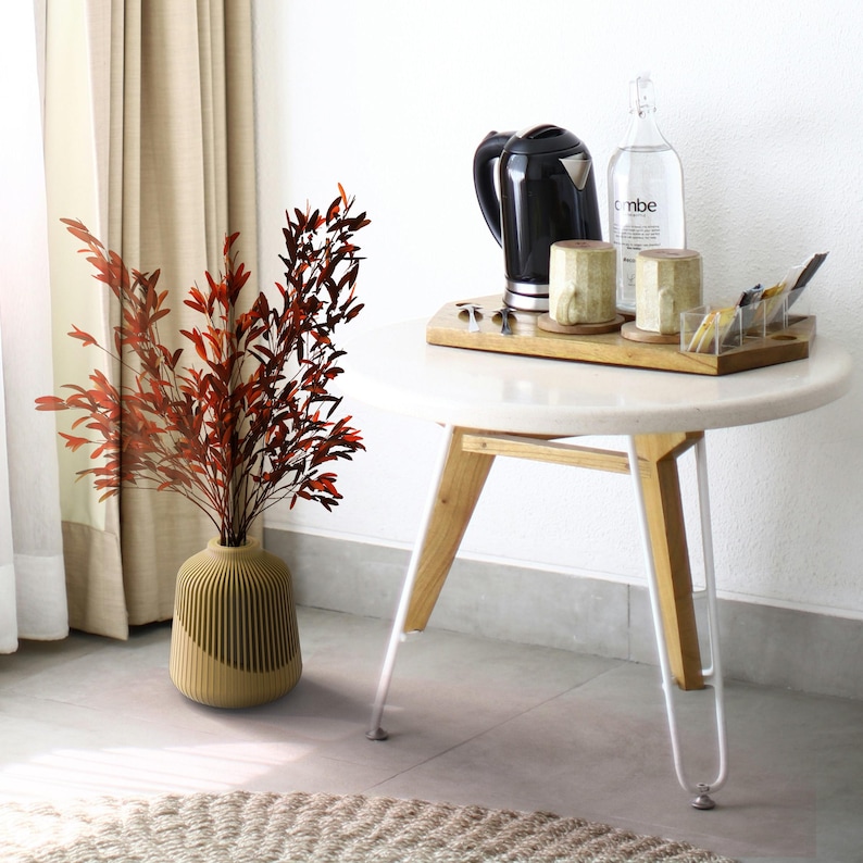 May include: A brown vase with a textured surface holds a bouquet of dried red leaves. The vase is on a light brown rug in front of a white table with a wooden top and white legs. On the table is a black electric kettle, two mugs, a glass bottle with the word "vibe" on it, and a wooden tray with various items.