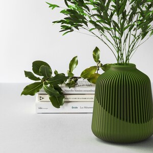 May include: A green ceramic vase with vertical lines, holding a bouquet of green leaves. The vase is sitting on a stack of books with French titles.