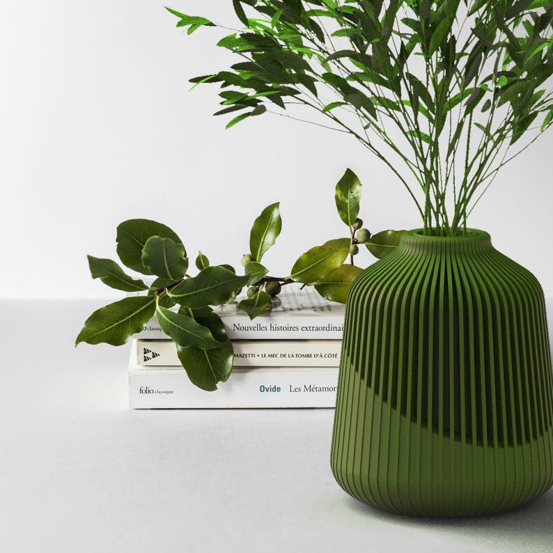 May include: A green ceramic vase with vertical lines, holding a bouquet of green leaves. The vase is sitting on a stack of books with French titles.