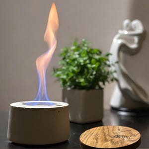 May include: A white concrete tabletop fireplace with a blue and orange flame burning inside. The fireplace is sitting on a wooden surface next to a potted plant and a white ceramic sculpture.