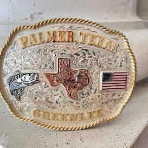 May include: Silver-toned metal belt buckle with gold-colored trim. The buckle features the text "PALMER, TEXAS" and "GREENVILLE" in gold lettering. It includes a Texas-shaped design, a fish, and an American flag.