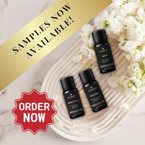 May include: Three small glass bottles with black labels and gold lettering. The bottles contain essential oil blends labeled "Nightfall", "Ascend", and "WFH". The bottles are arranged on a white, textured surface with white flowers in the background. The text "SAMPLES NOW AVAILABLE!" is displayed in gold lettering on a gold ribbon. The text "ORDER NOW" is displayed in red lettering on a red starburst.