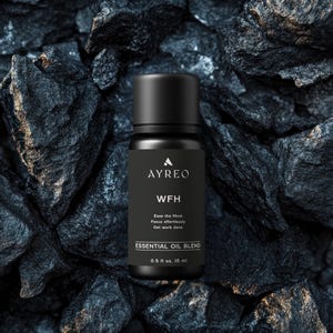 May include: A black essential oil blend bottle with the text "AYREO WFH Ease the Mind. Focus effortlessly. Get work done ESSENTIAL OIL BLEND 0.5 fl oz. 15 ml" on the label. The bottle is sitting on a bed of black rocks.