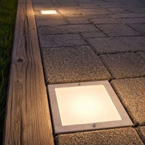 May include: A series of square, flush-mounted outdoor lights embedded into a gray stone paver walkway. Each light features a metallic frame with visible screws and emits a warm, soft glow, illuminating the textured surface of the stone path.