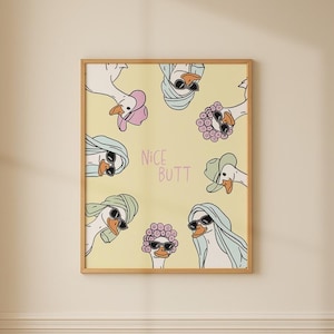 May include: A framed print featuring cartoon geese wearing sunglasses and various head coverings, set against a yellow background. The phrase "NICE BUTT" is written in the center. The artwork is displayed in a bathroom setting.