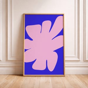 Könnte beinhalten: Gerahmter Kunstdruck mit einer großen, abstrakten rosa Form vor einem leuchtend blauen Hintergrund. Das Kunstwerk ist in einem dünnen, natürlichen Holzrahmen eingefasst. Die Gesamtästhetik ist modern und minimalistisch, passend für die Inneneinrichtung.
