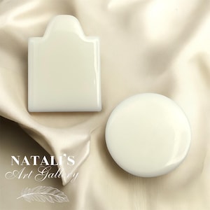 May include: Two white ceramic soap dishes, one shaped like a rectangle with a rounded top and the other a round disc. Both dishes are on a beige fabric background.