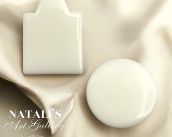 May include: Two white ceramic soap dishes, one shaped like a rectangle with a rounded top and the other a round disc. Both dishes are on a beige fabric background.