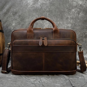 Puede incluir: Un maletín de cuero marrón con un asa superior y una correa para el hombro desmontable. La bolsa tiene un bolsillo frontal con cremallera y dos tiradores de cremallera más pequeños. El cuero tiene un aspecto envejecido.