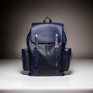 May include: A two-tone leather backpack in navy blue and black. The backpack features a flap closure with two buckle straps, side pockets, and a top handle. The bag is designed with a drawstring closure and silver-tone hardware.