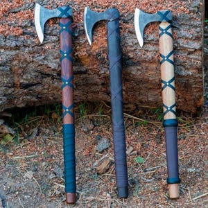 Op de afbeelding: Drie bijlen met zilverkleurige metalen koppen en houten stelen. Elke steel is voorzien van een kruislings lederen wikkeling vastgezet met metalen klinknagels en een zwarte lederen grip aan de basis. De stelen variëren in kleur van donker- tot lichtbruin.
