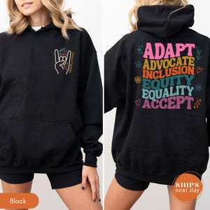 May include: Black hoodie with a front pocket and a colorful hand gesture graphic with the words "Social Education." The back of the hoodie features the words "ADAPT ADVOCATE INCLUSION EQUITY EQUALITY ACCEPT" in various colors.