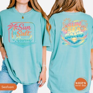 May include: A seafoam green t-shirt featuring a front graphic with a palm tree and the text Sun Salt Sand in a gradient of pink, orange, and blue. The back graphic displays a sunset over the ocean with palm trees and the text Sun Sea Sand.