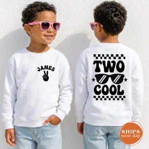 May include: A white long-sleeve sweatshirt featuring black text and graphics. The front displays the name James above a peace sign hand gesture. The back features a checkered border, the text Two Cool, and a graphic of sunglasses with stars.