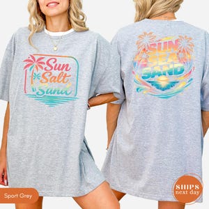 May include: A sport grey t-shirt featuring a front graphic with a palm tree and the text Sun Salt Sand in a square frame. The back graphic displays a colorful sunset, palm trees, and the text Sun Sea Sand over a stylized ocean wave design.