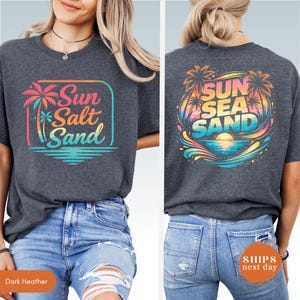 May include: A dark heather gray t-shirt featuring a front graphic with a palm tree and the text Sun Salt Sand in a gradient of pink, orange, and blue. The back graphic displays palm trees, a sunset, and the text Sun Sea Sand in vibrant colors.