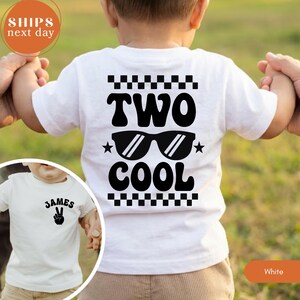 May include: A white short-sleeved t-shirt featuring a black graphic on the back with a checkered border, the text TWO COOL, and sunglasses. The front of the shirt displays the name JAMES above a black hand gesture graphic showing a peace sign.