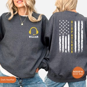 May include: A dark heather gray crewneck sweatshirt featuring a yellow headset icon and the name WILLIAM printed on the front left chest. The back displays a distressed white American flag graphic with the word DISPATCHER printed in yellow.