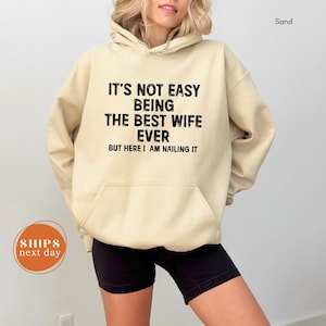 May include: Sand-colored hoodie with the text "IT'S NOT EASY BEING THE BEST WIFE EVER BUT HERE I AM NAILING IT" in black. The hoodie has a front pocket and a drawstring hood. The model is wearing black shorts.