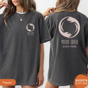 May include: A charcoal gray short-sleeve t-shirt featuring a light pink circular graphic of two hands reaching toward each other. The front chest displays a small version of the graphic with the text YOUR LOGO GOES HERE below it.