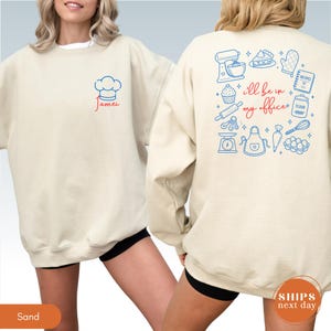 May include: A sand-colored crewneck sweatshirt featuring a blue chef hat and the red name Jamei on the front. The back displays blue baking icons including a mixer, cupcake, and scale, with the red text I'll be in my office.