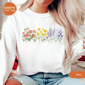 May include: White sweatshirt featuring a colorful floral design across the chest. The design includes pink, yellow, purple, and blue flowers. The sweatshirt has a crew neck and long sleeves. The text "Ships Next Day" is in a circle in the upper left corner.