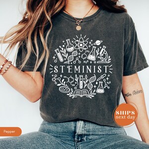 May include: A charcoal gray short-sleeve t-shirt featuring a white circular graphic print of various scientific icons including a microscope, DNA strands, beakers, and the word STEMINIST centered in the middle of the design.