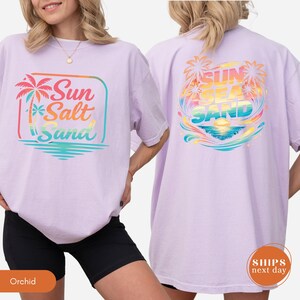May include: A light purple short-sleeve t-shirt featuring a graphic on the front and back. The front graphic shows a palm tree and the text Sun Salt Sand. The back graphic displays palm trees, a sunset, and the text SUN SEA SAND.