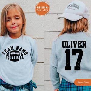 May include: A light grey crewneck sweatshirt featuring a black graphic of a football with the text TEAM NAME FOOTBALL on the front and the name OLIVER with the number 17 in black on the back, paired with a white Los Angeles baseball cap.