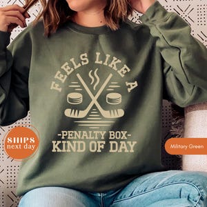 May include: Military green sweatshirt with a hockey-themed graphic. The design features crossed hockey sticks, pucks, and the text "FEELS LIKE A -PENALTY BOX- KIND OF DAY". The sweatshirt is a solid color.
