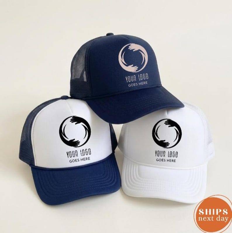 May include: Three trucker hats featuring a mesh back and a front panel with a circular logo of two hands forming a ring. One hat is navy blue with a light pink logo, while two are white with black logos. Each hat displays the text YOUR LOGO GOES HERE.
