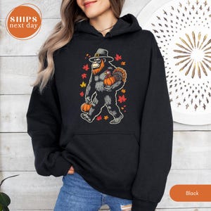 May include: A black hooded sweatshirt featuring a graphic of a Bigfoot creature wearing a pilgrim hat, holding a pumpkin and a turkey. The design is surrounded by scattered red and orange autumn leaves, set against a solid black fabric background.
