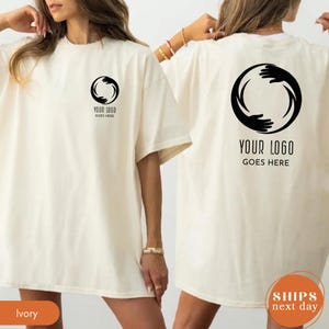 May include: An ivory t-shirt featuring a black circular graphic of hands on the left chest and a larger version of the same graphic on the back. The text "YOUR LOGO GOES HERE" is printed in black below both the small and large circular hand designs.