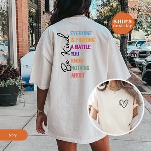 May include: Ivory-colored t-shirt with the words "Be Kind" and a message in multiple colors. The text on the back reads "Everyone is fighting a battle you know nothing about." A small heart design is on the front. The shirt is a casual, oversized fit.