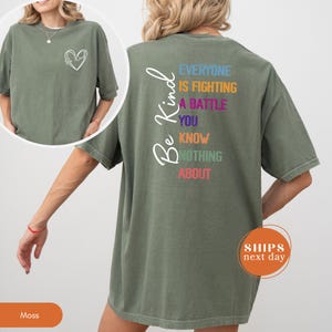 May include: Moss green t-shirt with a white heart outline on the front and the words "Be Kind" on the back. The back of the shirt also has the text "Everyone is fighting a battle you know nothing about" in various colors.