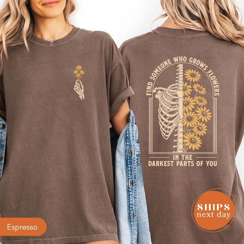a woman wearing a brown t-shirt with a graphic design on the back. The design features a skeleton with yellow flowers, and the text on the back reads "Find someone who grows flowers in the darkest parts of you".