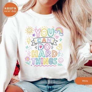May include: A white long-sleeve sweatshirt featuring a colorful graphic print with the text "YOU CAN DO HARD THINGS" in a crayon-like font. The design includes small illustrations of a sun, rainbow, flower, heart, and flexed arm muscle.