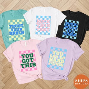 May include: Five short-sleeved t-shirts in various colors: teal, white, black, pink, and lavender. Each shirt features a checkered design with motivational phrases like "Trust Your Brain" and "You Got This" in bold text.
